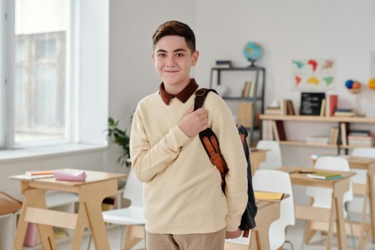 A smiling schoolboy confidently standing in a modern classroom with his backpack.